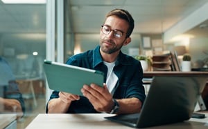 Professional reviewing a tablet at his desk with a laptop open in a modern office.